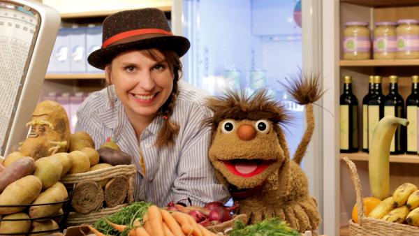 Maya und Moppi lachen hinter der Theke des Leckerladens. Vor Ihnen liegen Kartoffeln, Möhren, Zwiebeln und Bananen.